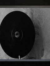 Czarno-biała grafika. Na pierwszym planie widać czarną okrągłą płytę z otworem w samym jej środku. Przez otwór spogląda ludzkie oko. W dalszej perspektywie widać, że za płytą stoi rezydentka.