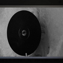 Czarno-biała grafika. Na pierwszym planie widać czarną okrągłą płytę z otworem w samym jej środku. Przez otwór spogląda ludzkie oko. W dalszej perspektywie widać, że za płytą stoi rezydentka.