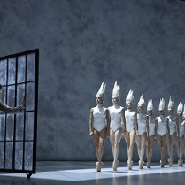 na zdjęciu po lewej stronie chłopiec patrzący przez duże okno, przed oknem rząd baletnic w białych strojach typu body i białych nakryciach głowy