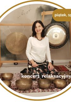 Kobieta w średnim wieku z czarnymi włosami. Ubrana w biały długi rękaw i wzorzyste spodnie. Siedzi na podłodze pokrytej dywanami. Za nią wiszą 2 gongi. Przed nią leży kilka mis.