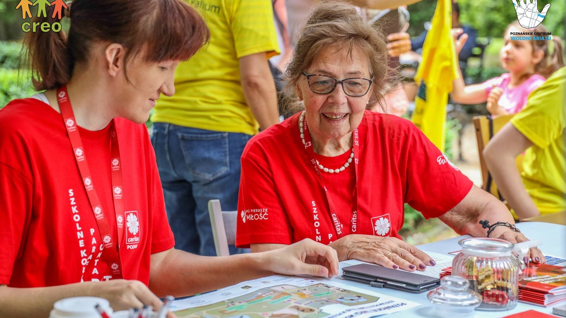 Na zdjęciu dwie wolontariuszki w czerwonych koszlkach siedzące przy stoliku, w tle dzieci