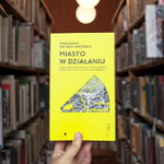 okładka książki na tle bibliotecznych regałów