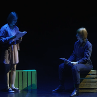Zdjęce dwóch osób na scenie czytających tekst, jedna osoba stoi, druga osoba siedzi na skrzynce.