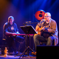 Zdjęcie przedstawia dwóch siedzących mężczyzn na scenie. Jeden z gitarą, drugi przy instrumencie klawiszowym.