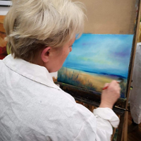 Starsza kobieta w białym fartuchu malująca obraz pędzlem na płótnie.