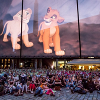 Grupa ludzi siedząca na ziemi pod ekranem z wyświetlonym filmem "Król lew".