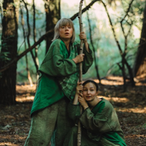 Dwie kobiety w lesie, ubrane w zielone atroje, trzymają cienki pień drzewa.