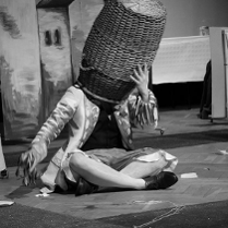 Czarno-białe zdjęcie osoby siedzącej na podłodze ze skrzyżowanymi nogami z dużym koszum na głowie; kosz całkowicie zakrywa twarz.