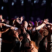 Pośrodku sceny stoi mężczyzna, wznosi ręce w górę, śmieje się, ciasno otacza go tłum ludzi, którzy starają się go dotknąć, śmieją się