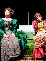 Dwie kobiety w długich, kolorowych sukniach, z bufiastymi rękawami siedzą ma scenie. W tle czarna ściana z białym zarysem drzwi.