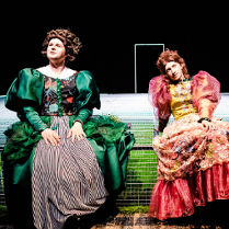 Dwie kobiety w długich, kolorowych sukniach, z bufiastymi rękawami siedzą ma scenie. W tle czarna ściana z białym zarysem drzwi.