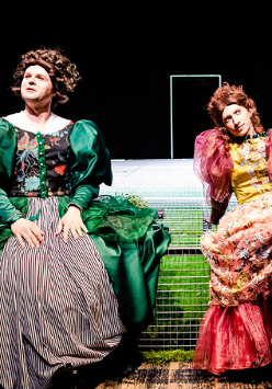 Dwie kobiety w długich, kolorowych sukniach, z bufiastymi rękawami siedzą ma scenie. W tle czarna ściana z białym zarysem drzwi.