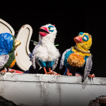 Na zdjęciu trzy kukuiełki kolorowych ptaków siedzacych na szarym murku.