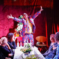 Przy stole siedzi kilka osób, u szczytu stołu stoi mężczyzna w kolorowym garniturze z szeroko rozłożonymi rękoma. Na stole kilka butelek alkoholu.