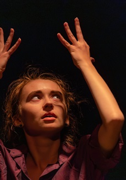 Kobieta w teatralnej pozie z uniesionymi rękoma do góry, ciemne tło.