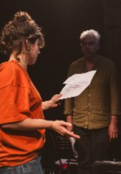 Kobieta oraz mężczyzna w półmroku na scenie. Kobieta jest zwrócona bokiem i w lewej ręce trzyma kartkę. Mężczyzna patrzy na kobietę na wprost.