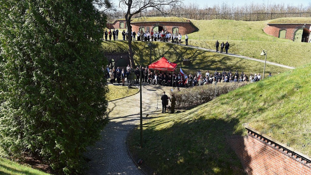 widok na zabudowania fortecze i pagórki porośnięte trawą, w centrum zdjęcia duża grupa ludzi biorących udział w uroczystościach