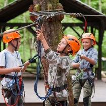 3 chłopców w kaskach w paku linowym.. W głębi stoliki, wiata, drzewa i krzewy.