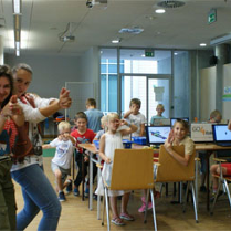 Dzieci siedza na krzesłach przy stolikach z laptopami. W sali sa instruktorzy.