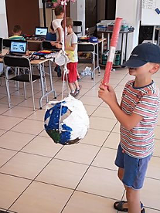 Sala, stoliki i krzesła. Na stołach laptopy. N apierwzym planie chłopiec z akrytymi czapka oczami z piankowa rurka próbuje trafić w zawieszona piłkę. Dalej 3 dzieci przy stolikach.