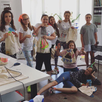 Zdjęcie grupowe w pracowni krawieckiej. Usmiechnietę Uczestniczki półkolonii z uszytymi workami sportowymi. Jedna osoba leży na podłodze.