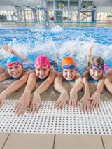 Grupa dzieci w czepkach w wodzie. Ręce mają wyciągnięte na brzegu basenu.