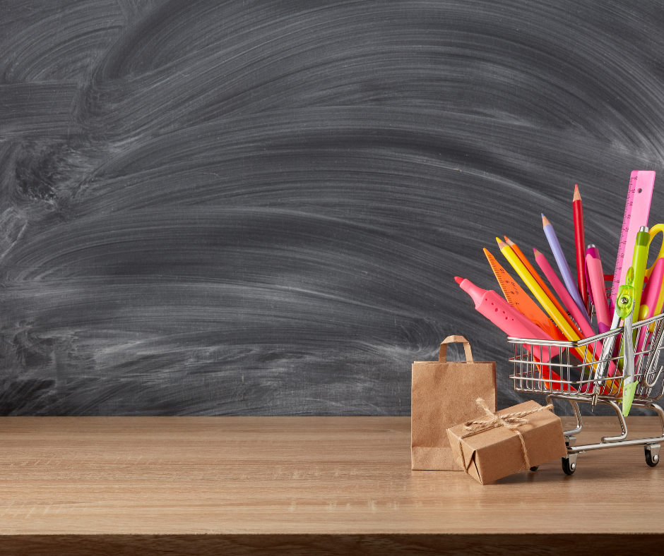 Na tle szkolnej tablicy znajduje się wózek sklepowy wypełniony przyborami szkolnymi