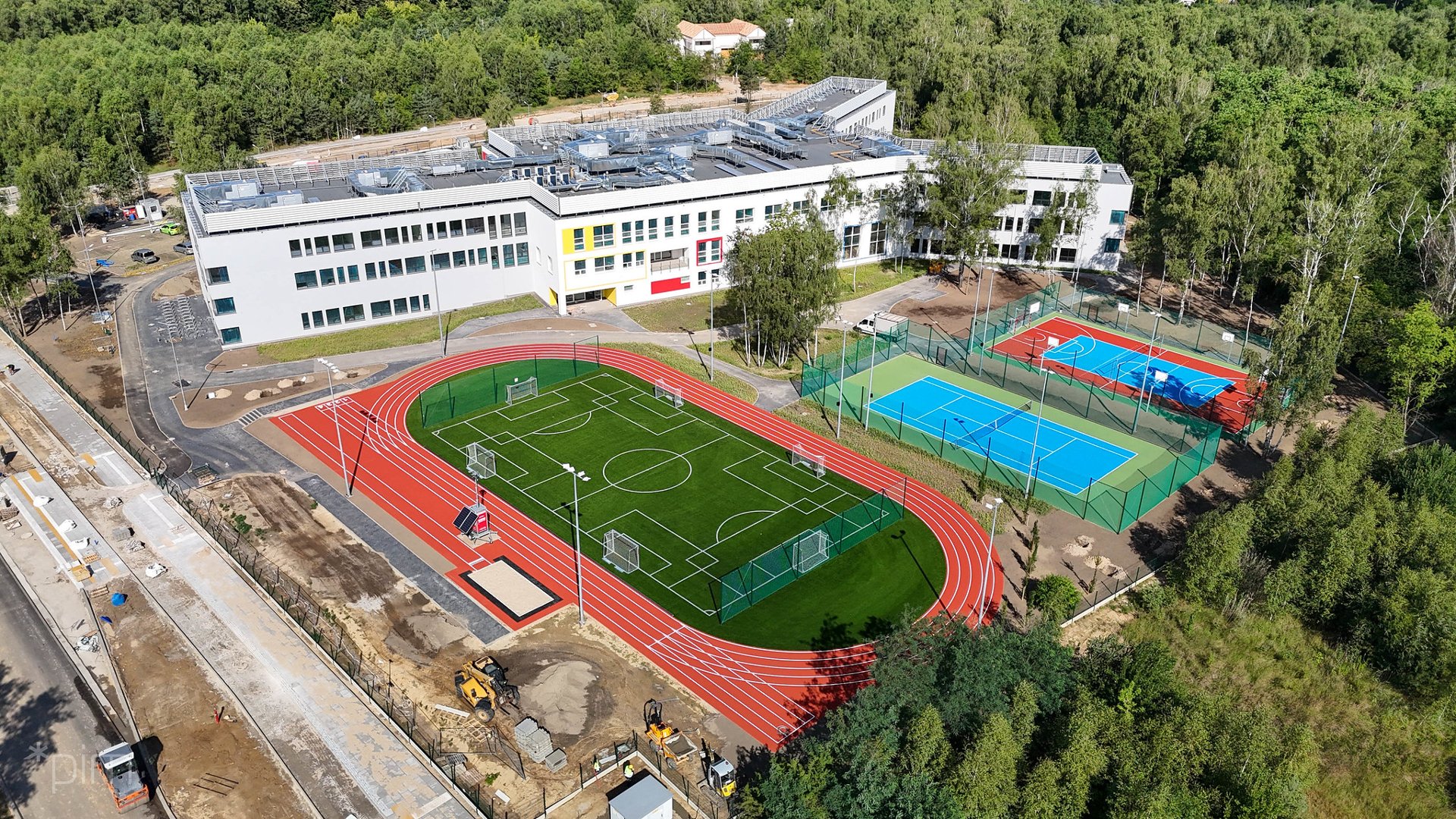 Na zdjęciu widać nowoczesny kompleks szkolny z lotu ptaka. Obok budynku znajdują się boiska sportowe - do piłki nożnej z bieżnią lekkoatletyczną, koszykówki i tenisa. Szkołę otacza zieleń i las.