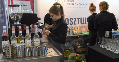 Zdjęcie przedstawia dziewczyny w czarnych koszulach na stanowisku gastronomicznym. Widać na nim takie akcesoria, jak szklanki, ekspres do kawy czy smakowe syropy do kawy w szklanych butelkach.