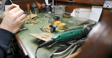 Na stole leżą fragmenty zegara i narzędzia zegarmistrzowskie.