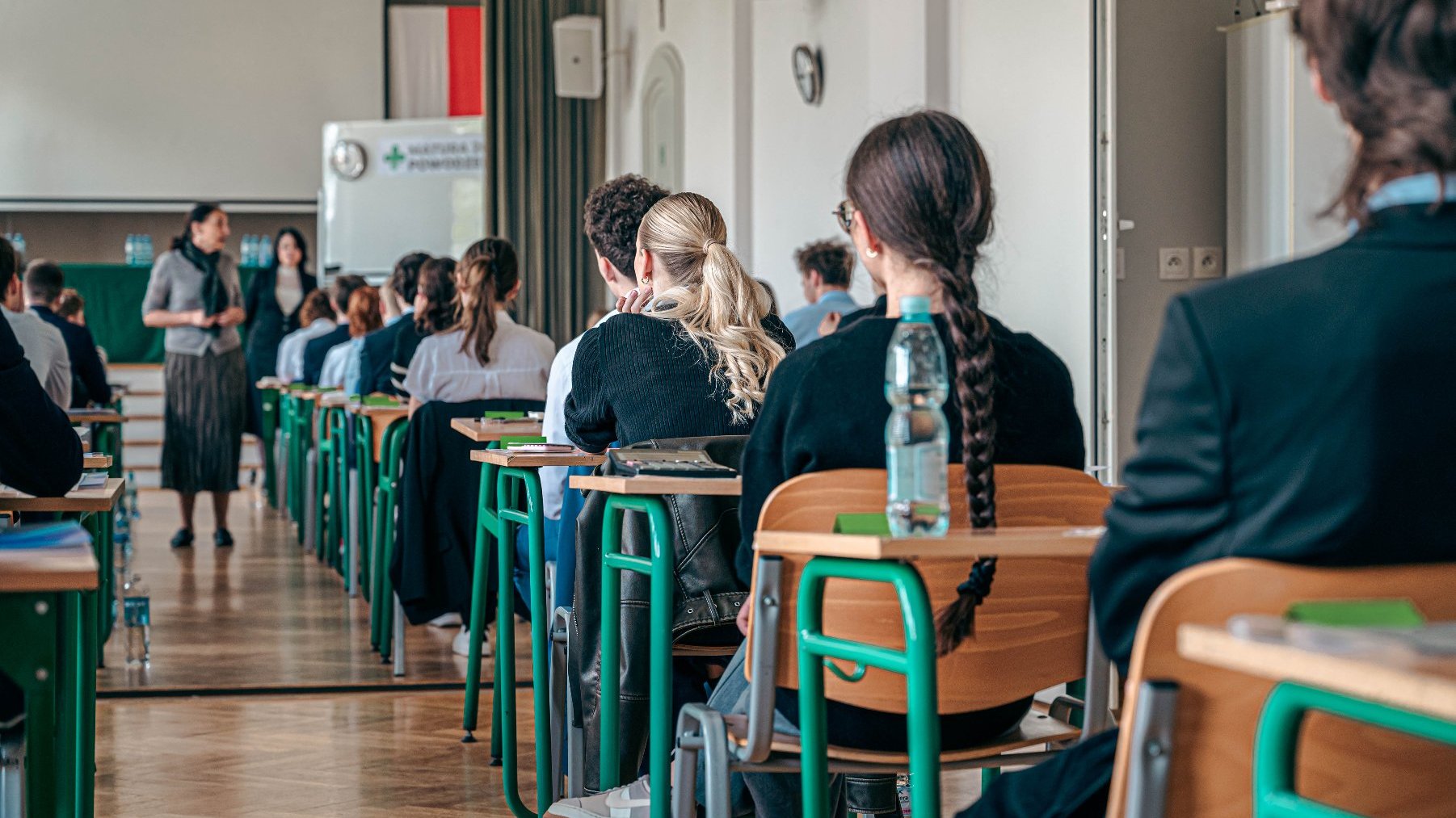 Zdjęcie przedstawia uczniów siedzących w ławkach na sali gimnastycznej, tyłem do aparatu.