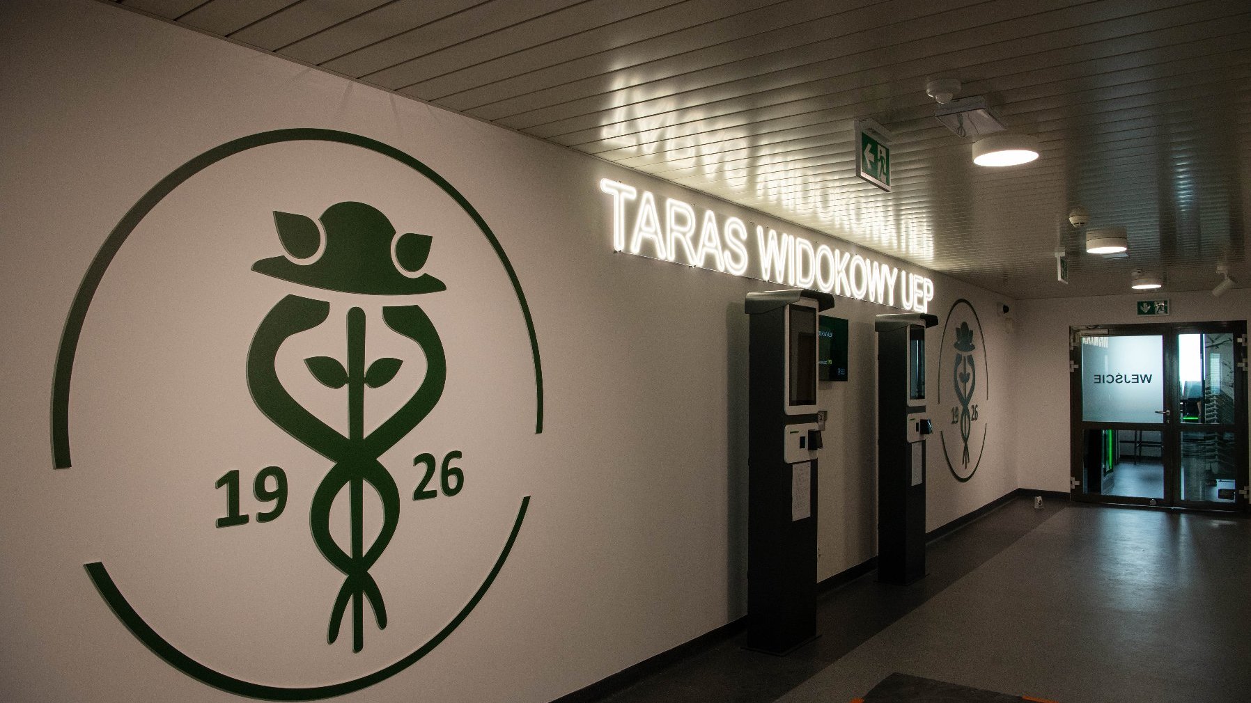 Korytarz z białymi ścianami. Na ścianie po lewej stronie znajduje się duże, zielone logo Uniwersytetu Ekonomicznego w Poznaniu z liczbami "19" i "26". Dalej, na ścianie, podświetlany napis "TARAS WIDOKOWY UEP". W oddali widoczne wejście do pomieszczenia.