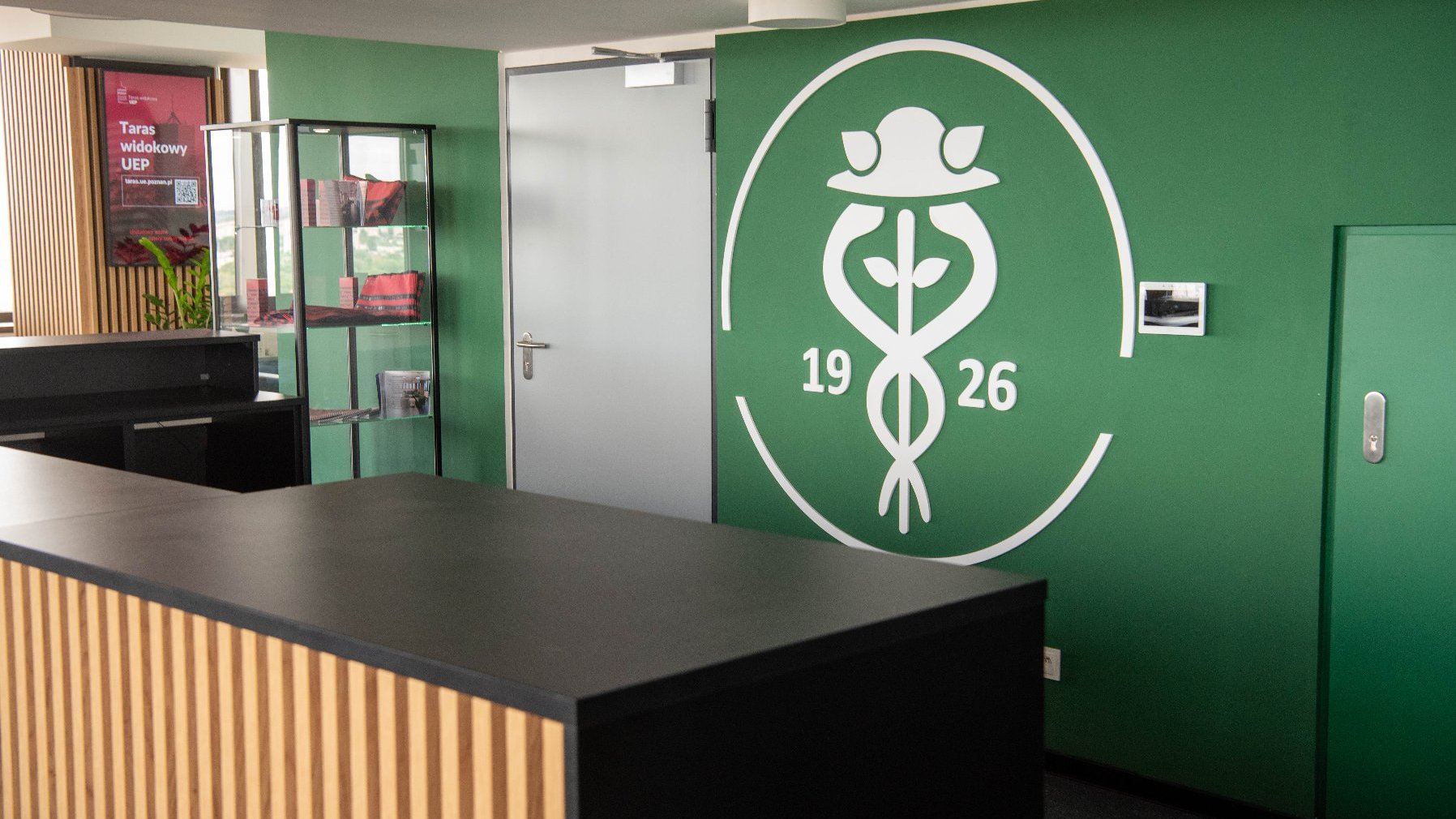 Wnętrze pomieszczenia, prawdopodobnie recepcja, z ciemnym blatem na pierwszym planie. Na zielonej ścianie widoczne jest duże, białe logo Uniwersytetu Ekonomicznego w Poznaniu z liczbami "19" i "26". Po lewej stronie znajduje się regał z materiałami promocyjnymi.