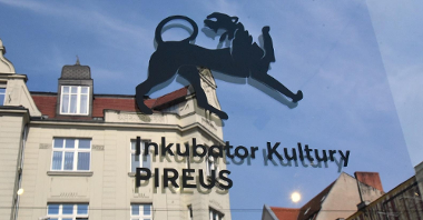 Zdjęcie przedstawia napis "Inkubator Kultury PIREUS umieszczony na szybie okiennej. Za oknem widać budynek kamienicy.