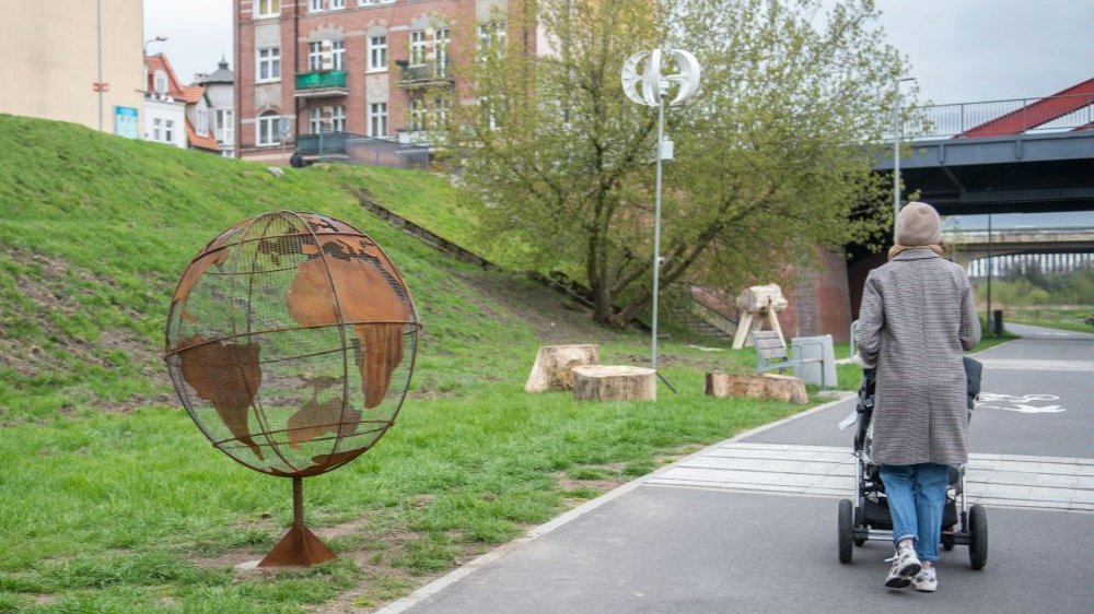 Zdjęcie przedstawia rzeźbę w kształcie kuli ziemskiej oraz instalacje pozwalającą ładować urządzenia dzięki sile wiatru. Obok widać kobietę z wózkiem. - grafika artykułu