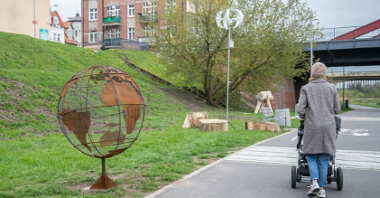 Zdjęcie przedstawia rzeźbę w kształcie kuli ziemskiej oraz instalacje pozwalającą ładować urządzenia dzięki sile wiatru. Obok widać kobietę z wózkiem.