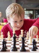 Chłopiec w czerwonej koszulce gra w szachy, za nim bawią się inne dzieci.