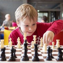 Chłopiec w czerwonej koszulce gra w szachy, za nim bawią się inne dzieci.