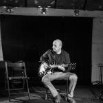 Zdjęcie mężczyzny siedzącego na krześle i grającego ma gitarze.