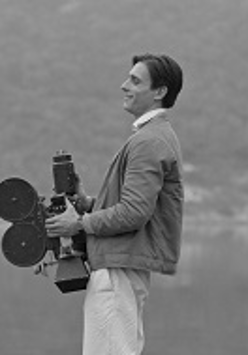 Czarno-białe zdjęcie mężczyzny stojacego bokiem do obiektywu i trzymającego kamerę na tle zamglonego krajobrazu.