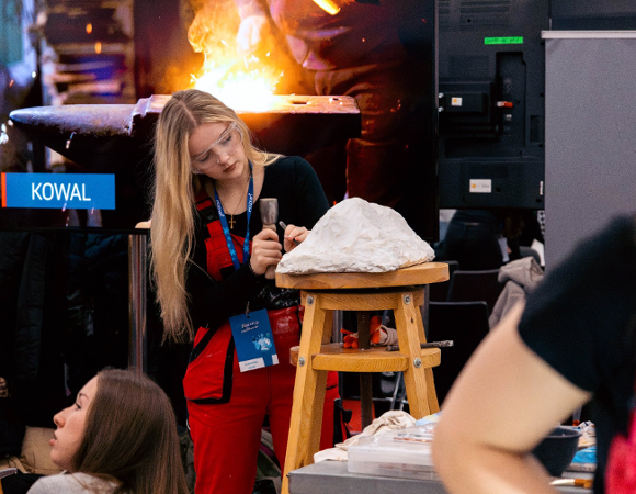 Zdjęcie przedstawia młodą dziewczynę rzeźbiącą w kamieniu stojącym na wysokim stołku.
