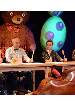 Na scenie czworo aktorów siedzi za stołem. W rękach trzymają uniesione drewniane łyżki, a na stołach leżą odwrócone do góry dnem garnki.