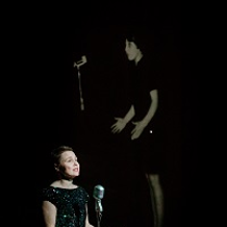 Kobieta w czarnej sukience stoi przed mikrofonem, za nią zdjęcie innej kobiety stojącej przed mikrofonem na czarnym tle.