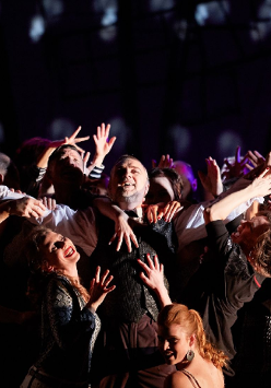 Pośrodku sceny stoi mężczyzna, wznosi ręce w górę, śmieje się, ciasno otacza go tłum ludzi, którzy starają się go dotknąć, śmieją się