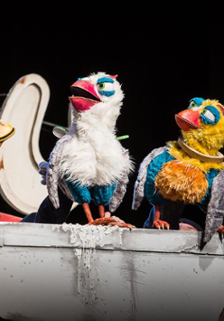 Na zdjęciu trzy kukuiełki kolorowych ptaków siedzacych na szarym murku.