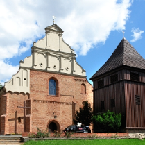 Zabytkowy kościół z cegły obok którego stoi drewniana dzwonnica. Fasada kościoła ma jasny szczyt. Dzwonnica ma ciemne, drewniane ściany. W tle widać niebo z białymi chmurami i drzewa otaczające budynki.
