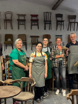 Grupa osób w fartuchach. Wokół przedmioty wykonane z drewna. W tle krzesła wiszące na szarej ścianie.