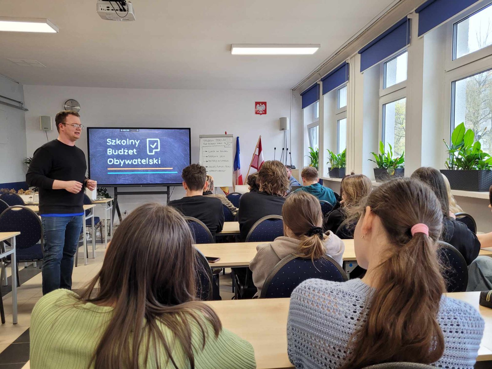 Grupa uczniów tyłem wraz z prowadzącym w klasie, w tle ekran z napisem "Szkolny Budżet Obywatelski" - grafika artykułu