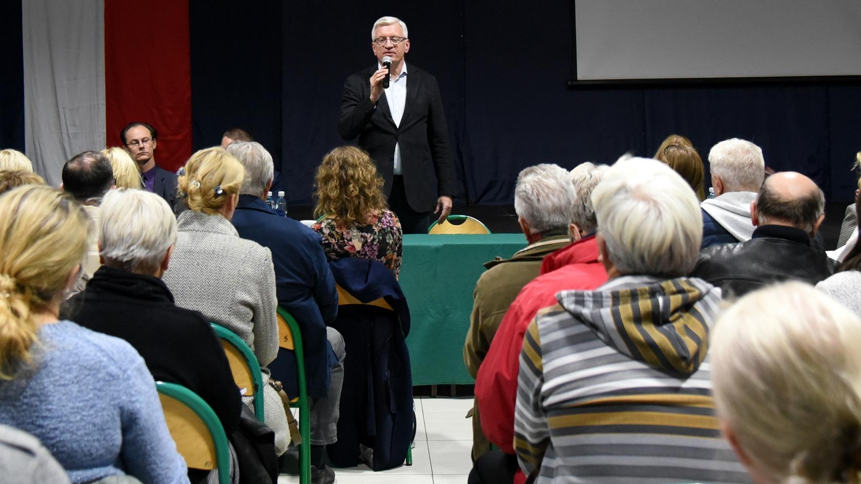 Zdjęcie przedstawia przemawiającego prezydenta Poznania, Jacka Jaśkowiaka. Prezydent stoi za stołem z mikrofonem, słucha go tłum ludzi. - grafika artykułu