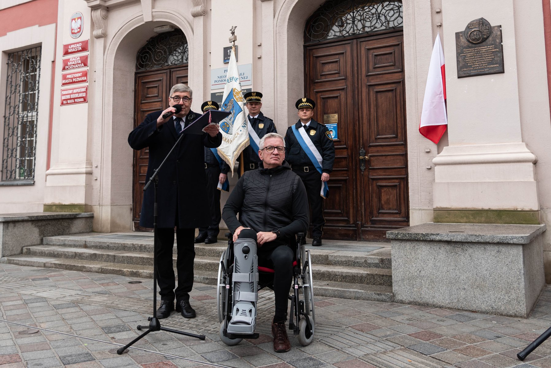 Na zdjęciu Jacek Jaśkowiak i Grzegorz Ganowicz przed wejściem do urzędu miasta, za nimi poczet straży miejskiej, na ścianie wisi odsłonięta tablica - grafika artykułu